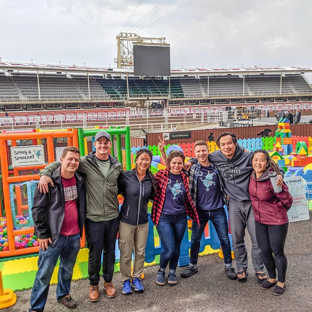 Seven people smiling, standing before a colorful playground and stadium seating.