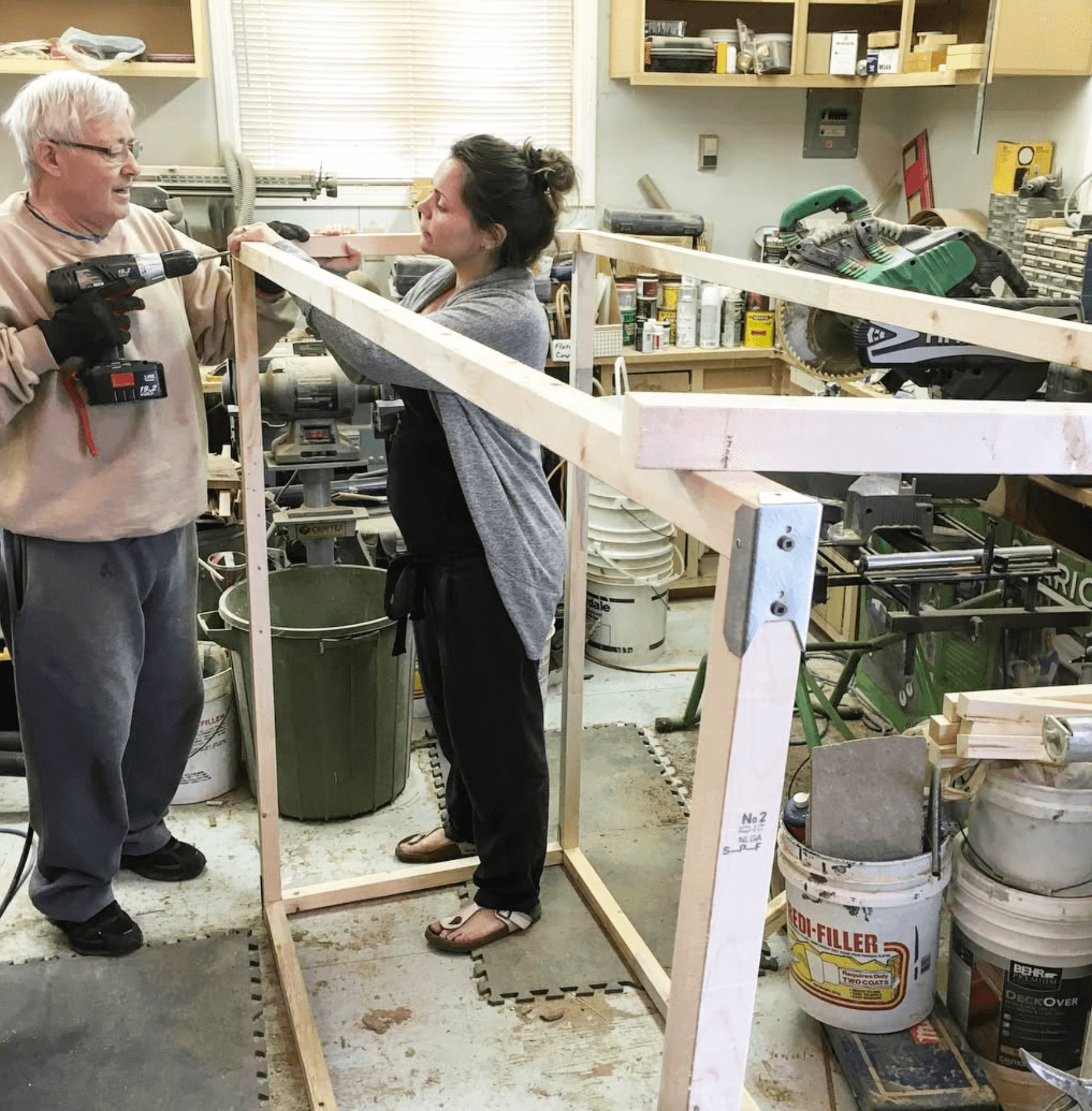 Man and woman building frame in cluttered workshop using tool and wooden beams.