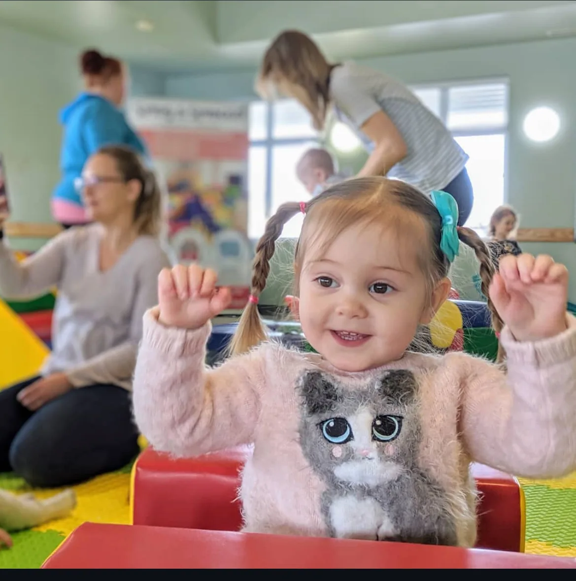 Smiling toddler with raised arms, surrounded by adults and children, colorfully dressed.