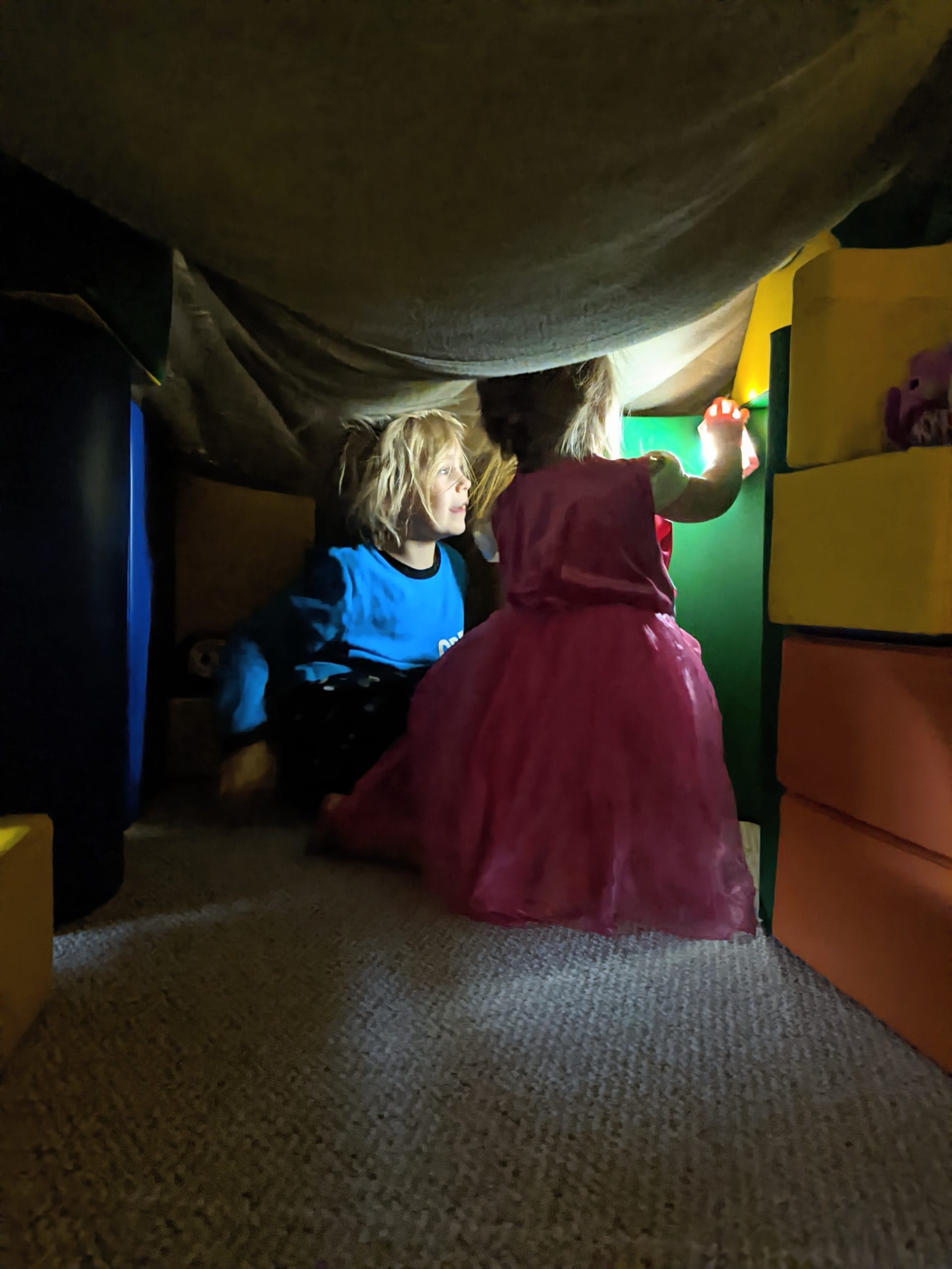 Two kids inside a blanket fort, surrounded by colorful cushions and soft light.