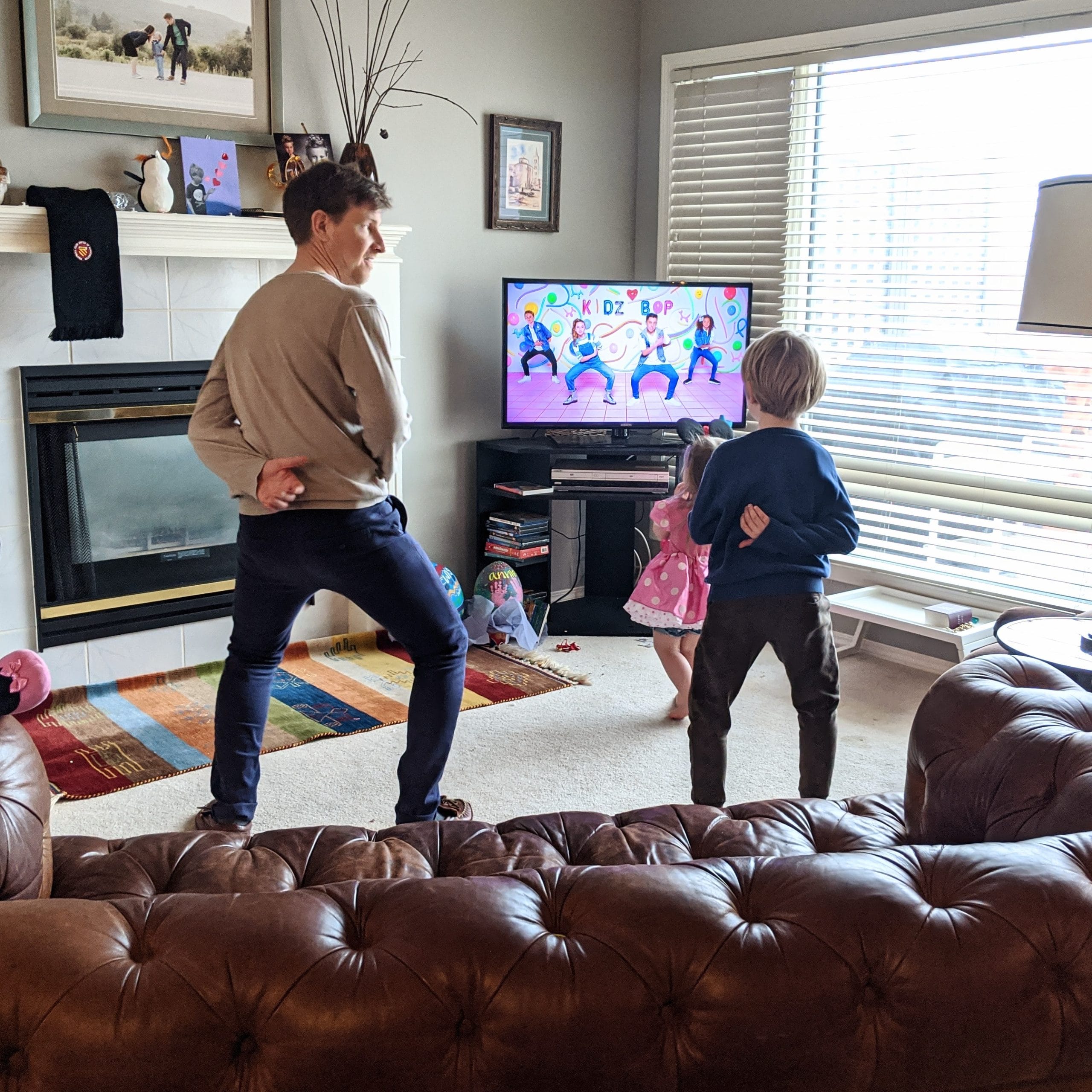 Two children and an adult dance in a living room with a TV on.