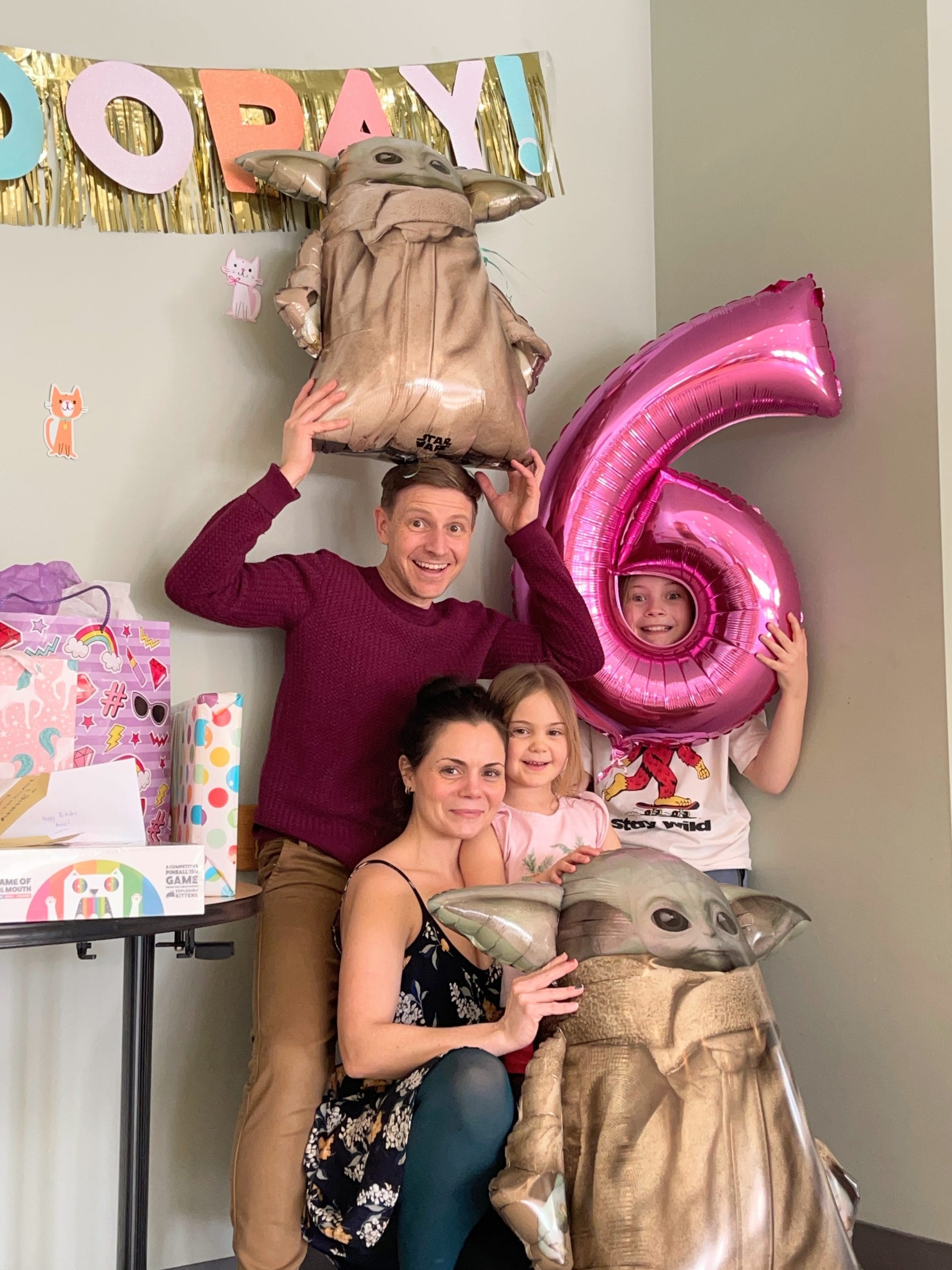 Family posing with large number six balloon and Grogu inflatables, surrounded by gifts.