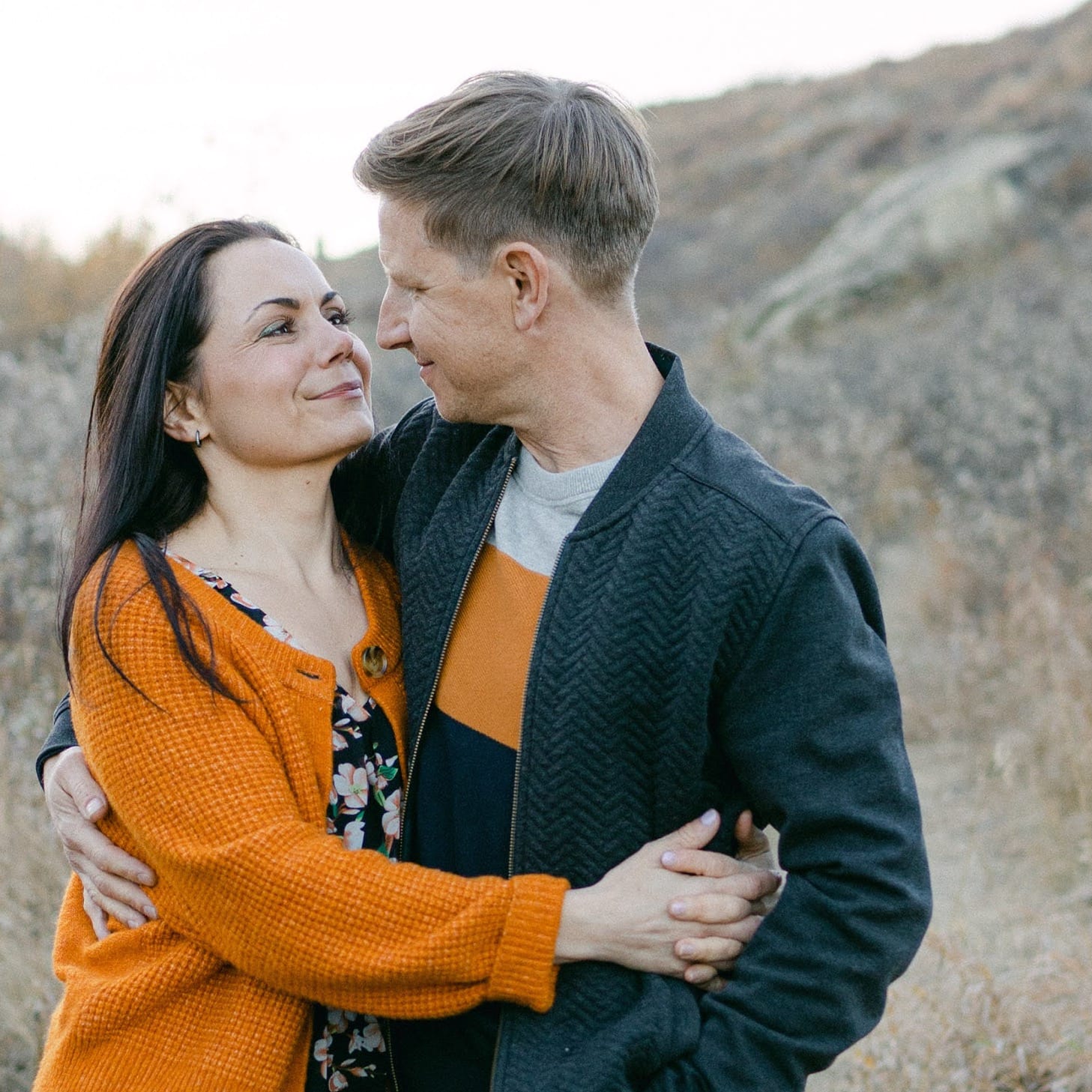 A couple in autumn attire embraces outdoors, with hills in the background.