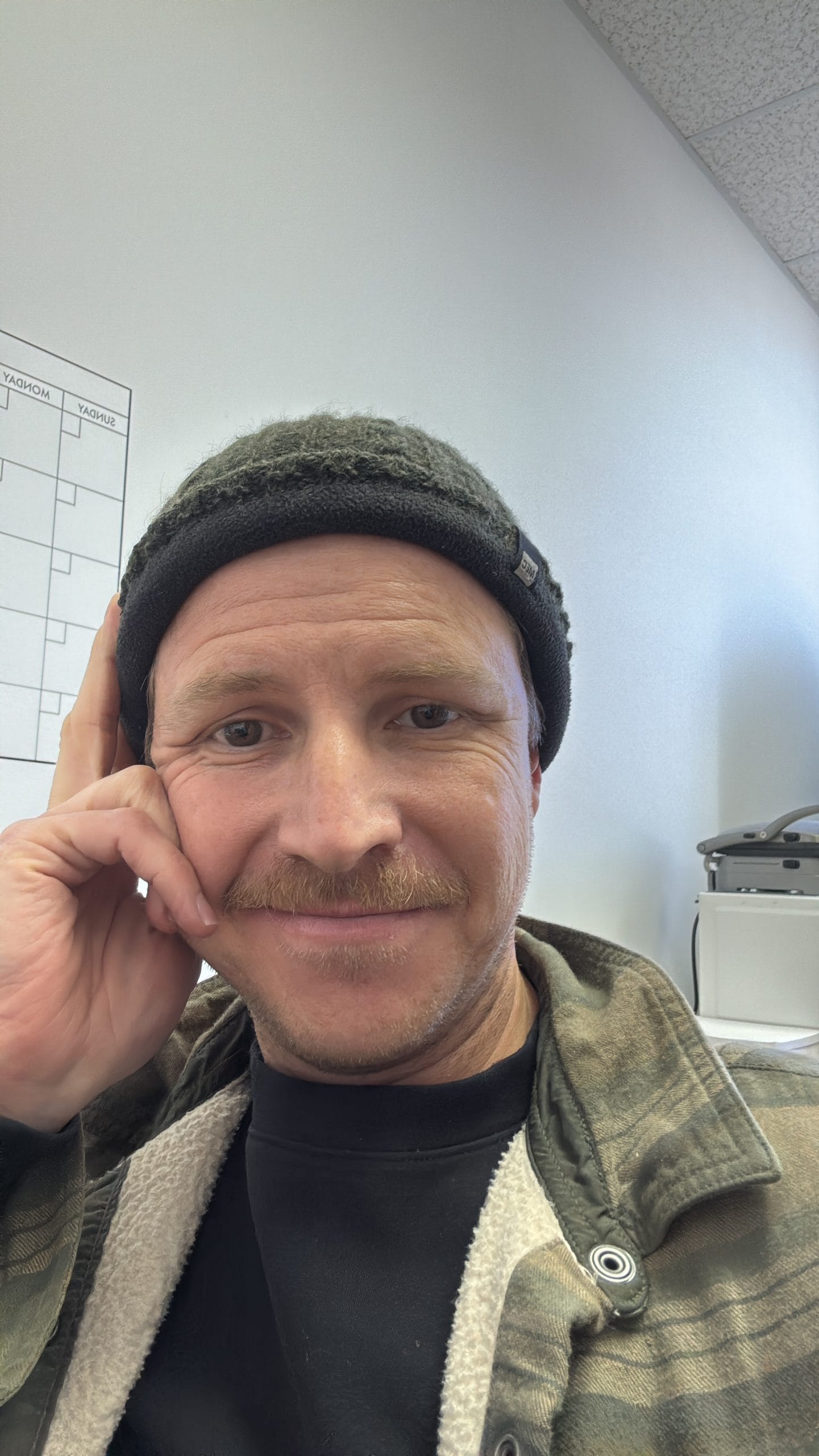 Man with a beanie and plaid jacket seated in an office environment.
